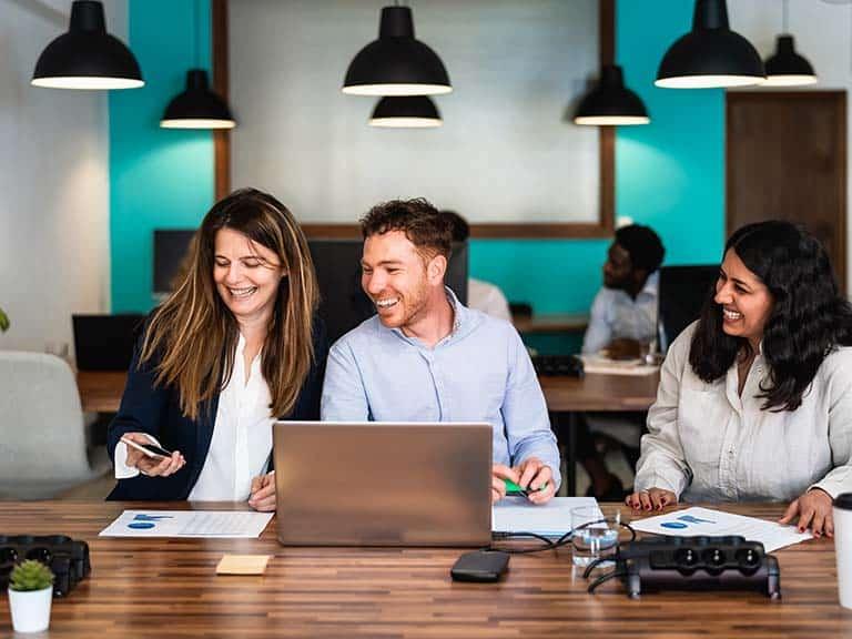 三位专业人士坐在一张桌子前，打开笔记本电脑，在一个共享的共同工作空间里工作，当一位女士拿起电话与另外两个人分享信息时，他们面带微笑.